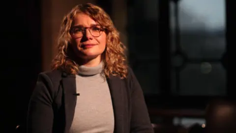 La dottoressa Kelly Johnson è seduta in una stanza. Ha lunghi capelli ricci e indossa una giacca sopra una giacca a collo alto e ha gli occhiali.