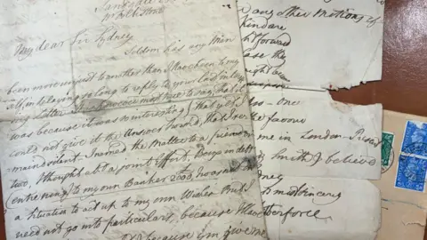 Hawleys Auctioneers The two letters with handwritten text on folded white paper on top of a manilla envelope with blue and green postage stamps. The letter on the left has several ink stains. The letter on the right is partially damaged with a wide tear halfway down. The handwriting is in neat lines and written with a flourish.