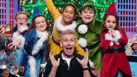 PA Media Patrick Kielty with five children behind him dressed as elves. He has blonde hair, wearing a navy shirt whit white t-shirt. The children are wearing elves costumes in either red, green, yellow or blue colours. A Christmas tree is behind them.