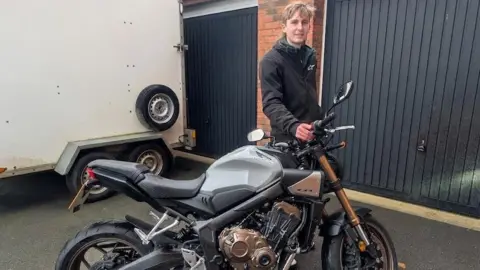 Gloucestershire Constabulary A young man stands on a driveway with his hand resting on the handlebars of a black and gold motorcycle. There are two dark painted garage doors behind him and a large white van beside him. He is wearing a black coat and has blond hair. He is smiling.
