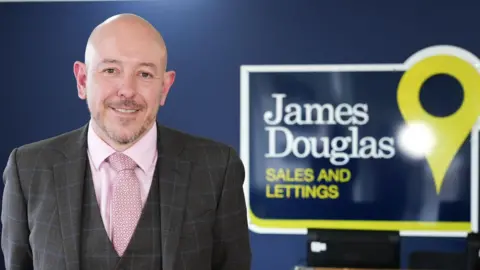 Douglas Haig stands in his estate agent office in front of a sign and next to a staircase. He is wearing a grey suit with a grey waistcoat, a pink shirt and tie and stands with his hands behind his back. He is smiling into camera. He is bald and has a little facial hair.