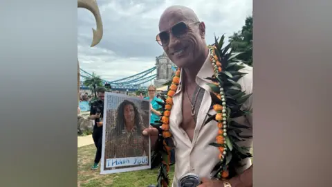 Jo Black/BBC The Rock – a man wearing a cream-coloured shirt and flowers around his neck. He is also wearing sunglasses and holding up a picture of himself as the character Maui. He is smiling.