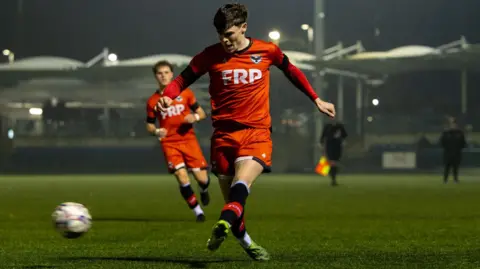 An FC IOM player kicks the ball while another player runs along behind him watching it.