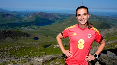 Nick Potts/PA Wire Olivia Clark is smiling at the camera. She is wearing a red football shirt, which has the Welsh dragon emblem to the right. Her hands are on her hips, and her brown hair is pulled back into a ponytail. She is standing on top of a mountain.