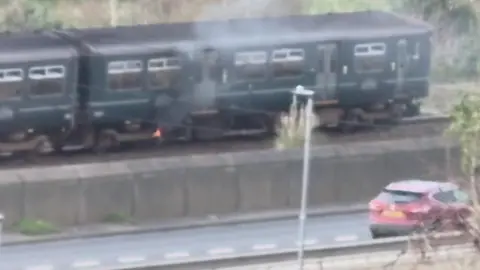 A train carriage has a fire underneath it with smoke emanating. In front of the train line is a road with one car driving past. The photo is hazy as if taken through a window.