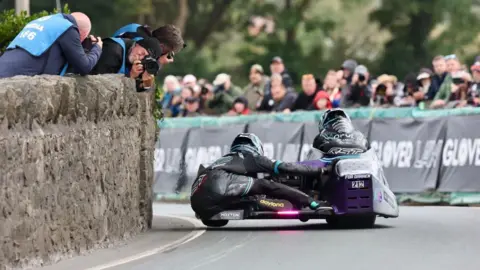 SOUTHERN 100 Sidecar outfit from behind as it rounds a stone wall bend. Spectators look on while photographers take pictures.