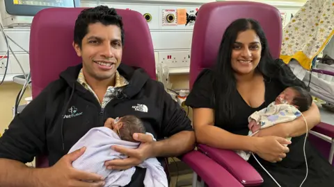 A man and a woman with dark hair each hold a tiny baby with feedings tubes coming from their noses