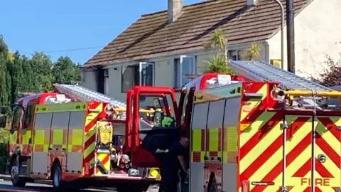 Two fire  engines outsdie a house with signs of smoke at a window.