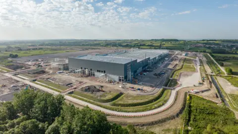 Agratas A large grey factory building under construction in the middle of a building site with trees and fields around stretching into the horizon it on a sunny day with blue skies and fluffy white clouds