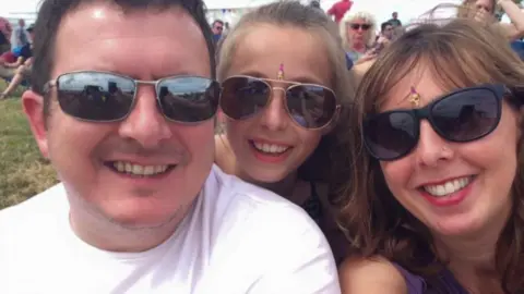Hollinson family Michael, Darcy and Debbie all smiling at the camera. They are all wearing sun glasses. There are people behind them , it looks like a festival.