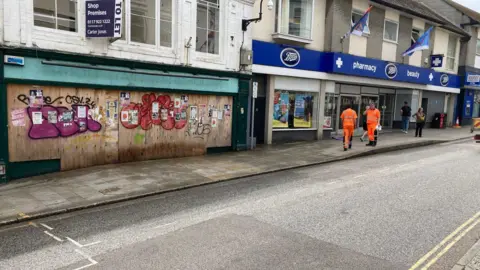 A wide shot of one of the shops on Market Jew Street which has been boarded up and covered with graffiti. There is a cleaning crew outside of it.
