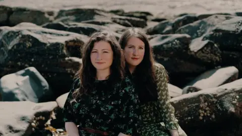 Supplied Rachel Unthank and Becky Unthank are sitting on a rocky beach with large stones behind them. One is sitting behind the other. They each have long dark hair and are wearing dark green tops 