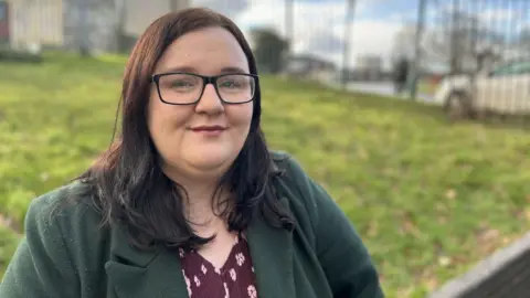 Labour councillor Hazel Dawkins is wearing a green coat, a red patterned top and glasses. She is smiling at the camera. The background is blurred but she is near a grassy verge on a road with a metal fence behind.