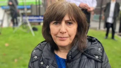 Gloria Levene with shoulder brown hair wearing a blue top and black coat standing outside the Heaton Park synagogue.