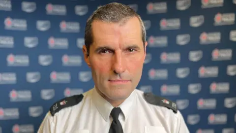 Stuart Woodward/BBC Leigh Norris is wearing a white shirt with black epaulettes on his shoulders and a black tie. He has short hair and a serious expression on his face. A blue banner with white Essex Police branding is behind him.
