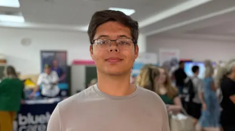 Wouter Le Roux has dark black hair and glasses and wears a light grey T-shirt. In the background are a number of out-of-focus people at a youth hub.
