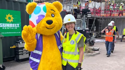 Claire Carrick Pudsey - a large yellow bear with a multi-coloured patch over part of his head - stands next to a woman on a building site. 
