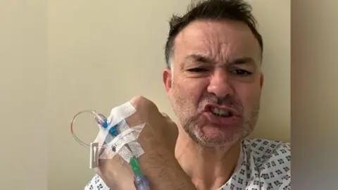 PA Media Lee Rawlinson is hospital holding his hand to the camera and pulling a face to show he is trying to be strong while undergoing treatment.