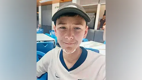 Mylo smiling into the camera. He is wearing a grey cap and England football shirt. He has a plaster on his chin.