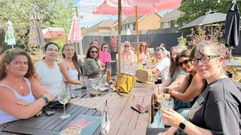 Contributed A large group of people seated around a long wooden outdoor table on a sunny day. Drinks, glasses, and small bags are spread across the table beneath colourful patio umbrellas. The gathering takes place in a garden-style pub setting with trees, fencing, and nearby houses in the background.