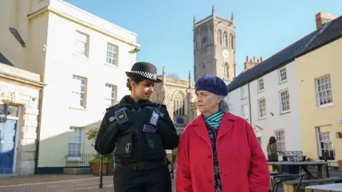 A still from the series showing Mandip Gill in character as a police officer standing next to Dawn French's character wearing a red coat and a beret. They are stood in a square with a church in the background.