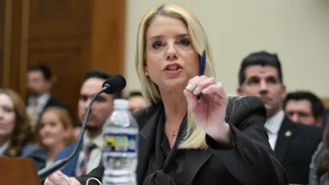 Pam Bondi gestures with pen in hand as she speaks into a microphone while sitting in a room in Capitol Hill with people in the background during a House Judiciary Committee hearing on "Oversight of the Department of Justice" on 11 February.
