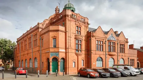 Historic England The exterior of Salford Lads and Girls Club. It is a red brick building with a green dome on top, and green doors.