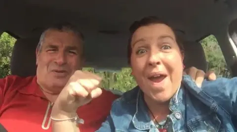 Barry Jones and Ali Alcock pictured in a still from a short video, with dad Barry smiling and Ms Alcock raising her fist in the air as they sit in the car. Ms Alcock is also smiling widely.