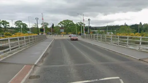 A red car is on the deck of Torvean Swing Bridge followed by another car. There are metal barriers on either side of the bridge.
