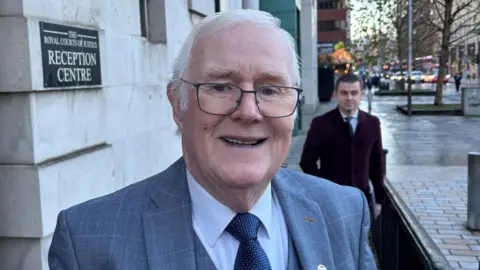 A close-up image of Eugene Reavey. He is an older man with medium-length white hair. He is wearing a pair of metal framed glasses and a grey checked suit jacket and waistcoat. He is also wearing a white shirt and a blue patterned tie. He is standing outside the Belfast High Court.