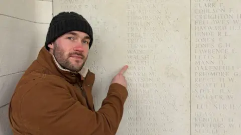 Philipp Cross Philipp wears a brown coat and black beanie hat and points to Percy Walsh's name engraved in stone