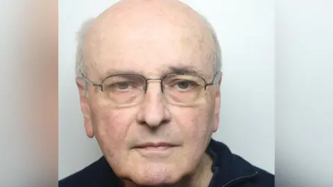 A mugshot of a man with white hair, a balding head and brown eyes. He is wearing spectacles and a navy jumper.