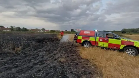 An all terrain fire brigade vehicle parked by a burnt area of grassland. A firefighter is hosing down undamaged grassland adjacent to the burnt area. 