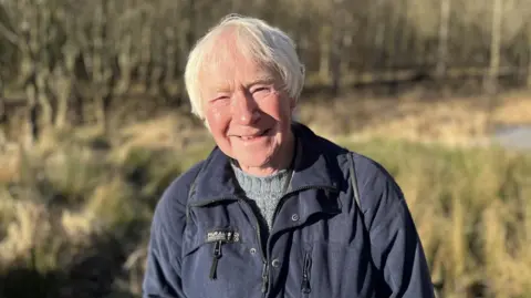 A grey-haired man with a dark blue jacket and grey jumper smiles at the camera, with a grassy area behind him