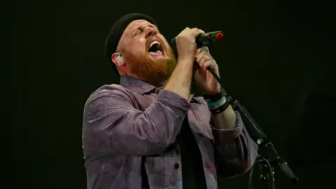 A wide shot of Tom Walker singing into a microphone. He is wearing a woolly hate and a jacket.