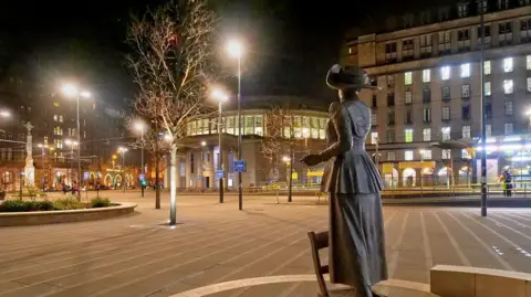 David Dixon/Geograph St Peter's Square at night with the statue of Emmeline Pankhurst in the foreground with the tram stop, town hall annexe and 