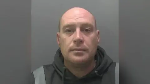 Custody photo of Darren Wyer, who is standing in front of a grey background. He has no hair and is looking directly at the camera. He wears a black hoodie with wide grey stripes on the shoulders. 