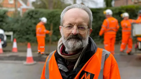 A grey-haired man with a grey beard and glasses, wearing a scarf and high-vis jacket, standing in front of road maintenance workers and traffic cones