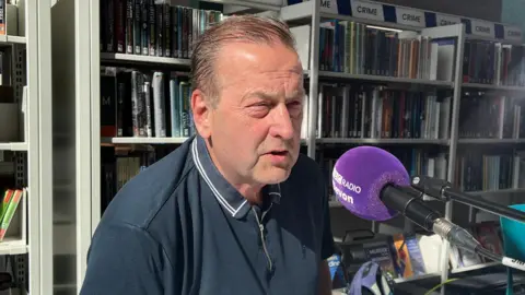 A man sitting in a library is talking into a purple microphone. A bookshelf is behind him. 