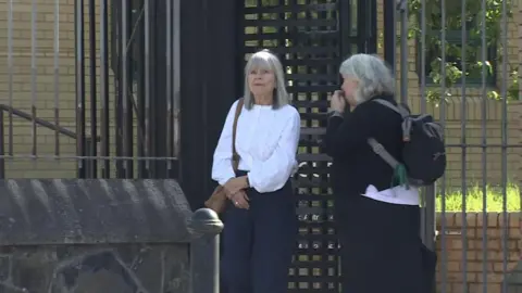 Two women leaving a black turntable style gate. The woman on the left is wearing a white blouse and navy trousers. She has a brown bag on her left shoulder and has straight grey hair to her shoulders and a fringe. The other woman is standing side on and has grey short hair and is all in black. Her face is obscured. She has a black backpack on. 