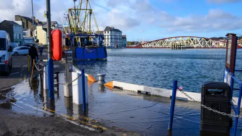 Manx Scenes Ramsey quayside at high tide, where water from the harbour has overflowed onto the pavement. There is a large blue fishing boat to the left and an arch steel bridge in red and white in the distance to the right.
