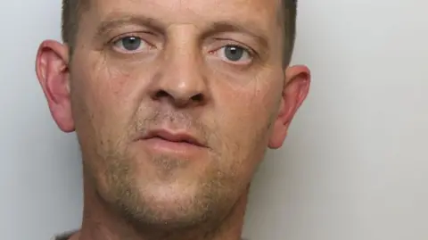 A police mugshot of a man aged around 40. He has receding brown hair, blue eyes and stubble. He is wearing a grey top.