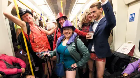 Getty Images Woman with no trousers wearing purple hat and holding blue suitcase while other passengers smile and a man with no trousers hold a coffee cup