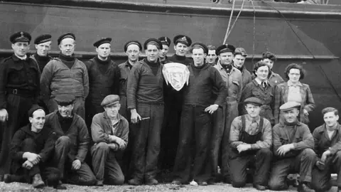 The Scalloway Museum A black and white picture of a group of about 18 men and two women. They stand shoulder to shoulder in a line, with some of the men on one knee in front of the group. Most wear navy caps. Lt David Howarth in the centre is holding the crest of the base: 'Alt for Norge'. 3rd right from Howarth (as viewed in the photo) is Severin Roald, Norwegian foreman of the slipway