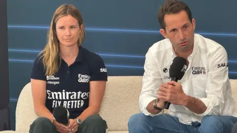 Hannah Mills wearing a blue GBR t-shirt and holding a microphone sat next to Sir Ben Ainslie who is wearing a white GBR shirt and talking into a microphone