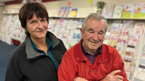 The photo shows Monika and Colin Morris standing in their shop, with shelves of greeting cards behind them.