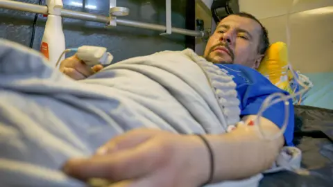 Bearded man lying in a hospital bed with a blue shirt and covered by a sheet. It joins a drip diet to your left arm. 