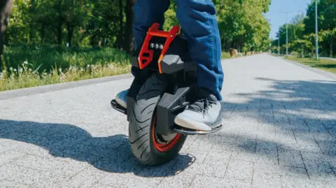 Getty Images Two legs on top of a red and black electric unicycle - a one-wheeled, electrically powered vehicle.