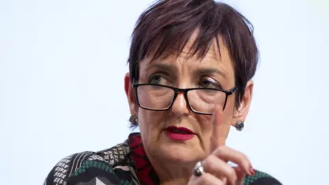 A woman with short dark hair, wearing glasses, in a close-up shot. She has her right hand raised in front of her face 