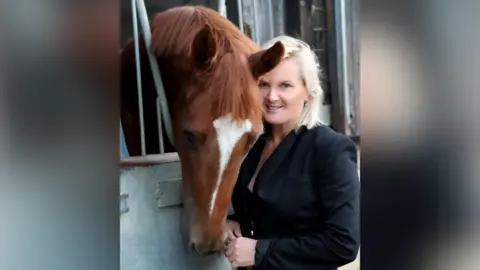 Contributed A picture of Gay Kelleway with a chestnut coloured horses. The horse has its head over the stable door and is looking down. Gay is smiling at the camera. She has short blonde hair and it wearing a black suit.